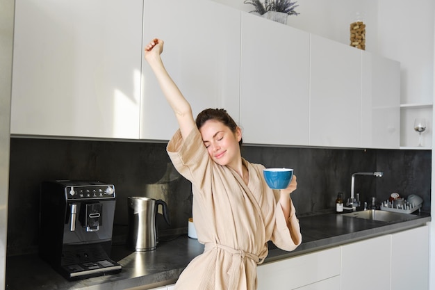 Portret szczęśliwej dziewczyny tańczącej z kawą w kuchni w szlafroku, cieszącej się porankiem