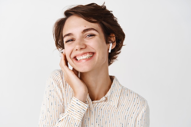 Portret szczęśliwej kobiety uśmiechającej się i słuchającej muzyki w słuchawkach, dotykającej słuchawek, stojącej na białej ścianie