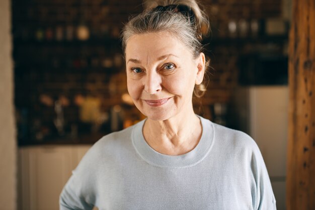 Portret szczęśliwej kobiety w średnim wieku ze zmarszczkami i niebieskimi oczami, która jest w dobrym, pozytywnym nastroju, spędzając miło czas w domu, pozując na tle przytulnej kuchni, patrząc na kamerę z wesołym uśmiechem