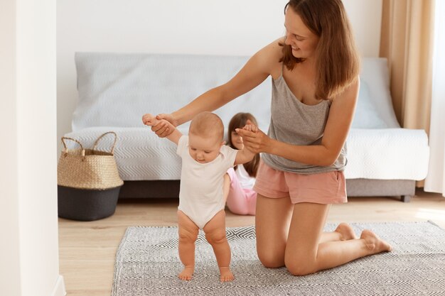 Portret szczęśliwej matki stojącej na kolanach na podłodze w salonie i uczącej swojego niemowlęcia, aby iść, maluch dziewczyna uczy się iść, szczęśliwe dzieciństwo.