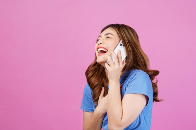 Portret szczęśliwej młodej kobiety noszącej zwykłą koszulkę rozmawiającą na telefonie komórkowym na białym tle na różowym tle