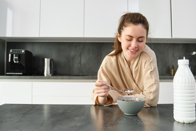 Portret szczęśliwej młodej kobiety opiera się na blacie kuchennym i je płatki zbożowe, ma przed sobą mleko i miskę
