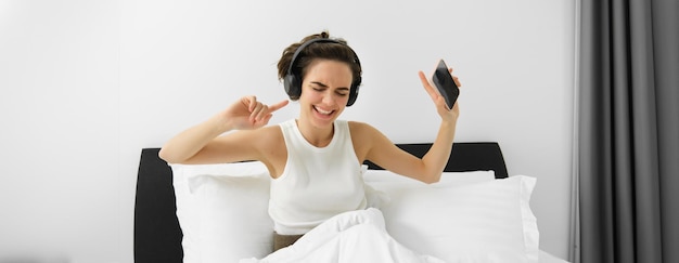 Bezpłatne zdjęcie portret szczęśliwej młodej kobiety w łóżku słuchającej muzyki w bezprzewodowych słuchawkach tańczących w niej