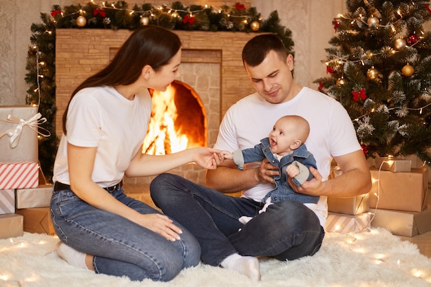 Portret szczęśliwej rodziny, ojca, matki i córki malucha pozuje przy kominku i choince w domu, ciesząc się, że możemy razem świętować ferie zimowe, szczęśliwego nowego roku.