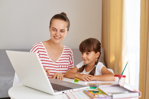 Portret szczęśliwej uśmiechniętej matki siedzącej obok córki małej uczennicy i odrabiającej pracę domową, kobieta pomagająca dziecku z lekcją online, mająca pozytywny wyraz.
