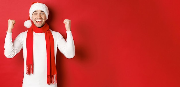 Portret szczęśliwy i podekscytowany mężczyzna w santa hat i szalik, radujący się i wygrywający coś, świętujący nowy rok, stojący na czerwonym tle.