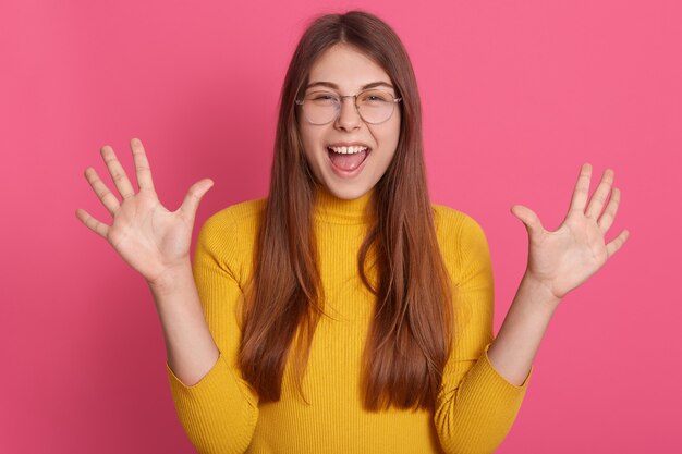 Portret szczęśliwy podekscytowana kobieta nosi żółty strój casual i okulary, podnosząc dłonie, wyrażając pozytywne emocje