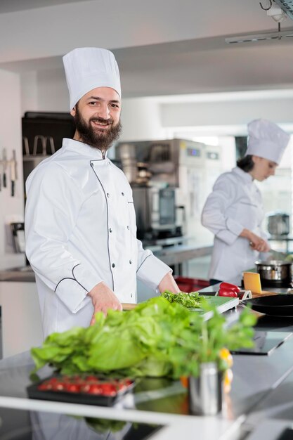 Portret szczęśliwy szef kuchni w mundurze do gotowania, stojąc w profesjonalnej kuchni restauracji, uśmiechając się do kamery. Wytrawny ekspert kuchni dla smakoszy przygotowujący składniki na danie.