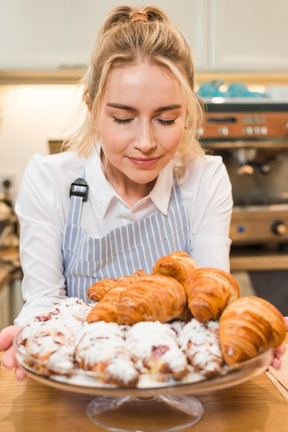 Bezpłatne zdjęcie portret szczęśliwy żeński piekarz bierze odory piec croissant w nim szklany torta stojak
