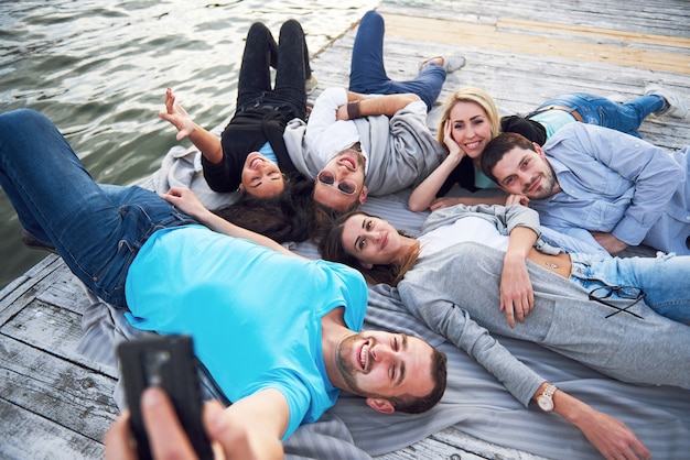 Bezpłatne zdjęcie portret szczęśliwych młodych przyjaciół na molo nad jeziorem. ciesząc się dniem i robiąc selfie.