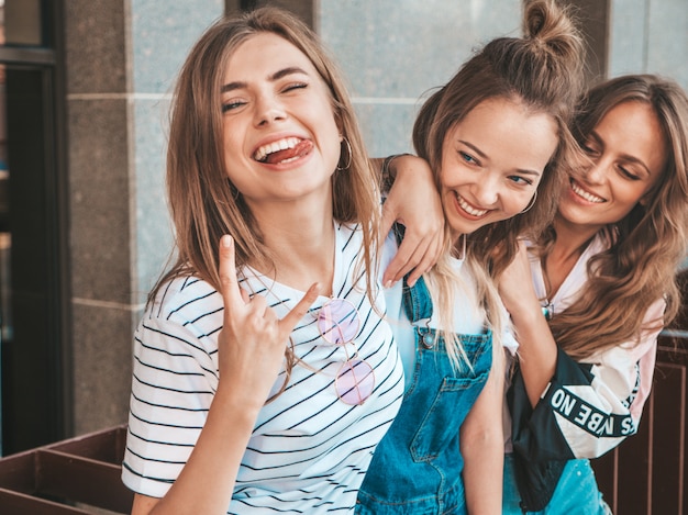 Bezpłatne zdjęcie portret trzech młodych pięknych uśmiechniętych hipster dziewcząt w modne letnie ubrania. seksowne beztroskie kobiety pozowanie na ulicy. pozytywne modele zabawy. pokazują język i rock and roll znak