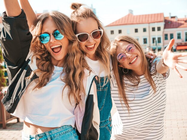 Portret trzech młodych pięknych uśmiechniętych hipster dziewcząt w modne letnie ubrania. Seksowne beztroskie kobiety pozowanie na ulicy. Pozytywne modele zabawy w okularach przeciwsłonecznych. Przytulanie