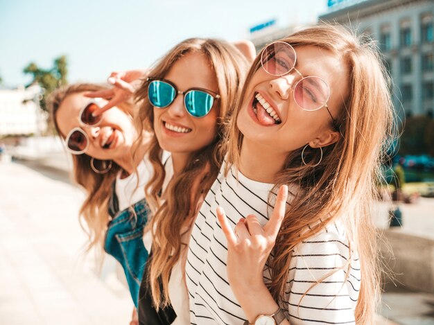 Portret trzech młodych pięknych uśmiechniętych hipster dziewcząt w modne letnie ubrania. Seksowne beztroskie kobiety pozowanie na ulicy. Pozytywne modele zabawy w okularach przeciwsłonecznych. Przytulanie