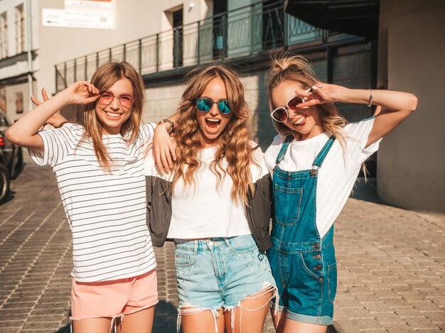 Portret trzech młodych pięknych uśmiechniętych hipster dziewcząt w modne letnie ubrania. Seksowne beztroskie kobiety pozowanie na ulicy. Pozytywne modele zabawy w okularach przeciwsłonecznych. Przytulanie