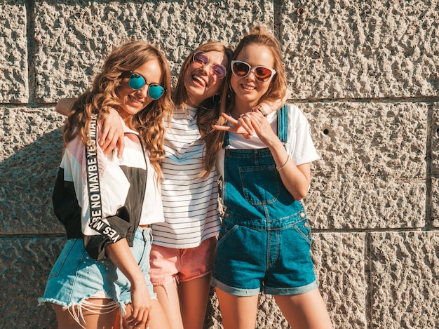 Portret trzech młodych pięknych uśmiechniętych hipster dziewcząt w modne letnie ubrania. Seksowne beztroskie kobiety pozuje w ulicznej pobliskiej ścianie. Pozytywni modele ma zabawę w okularach przeciwsłonecznych przytulenie