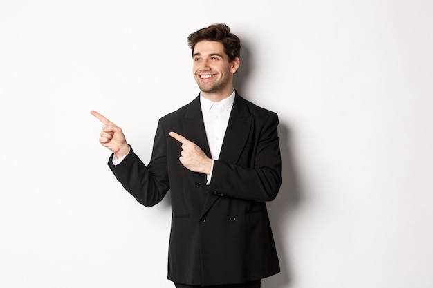 Portret udanego, przystojnego mężczyzny w garniturze, wskazującego i patrzącego w lewo z zadowolonym uśmiechem, pokazującym baner promocyjny, stojący na białym tle.