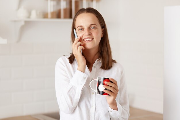 Portret ujmującej kobiety o ciemnych włosach rozmawia przez inteligentny telefon, trzymając kubek w ręce, ciesząc się kawą lub herbatą, mając przyjemną rozmowę, patrząc na kamery.