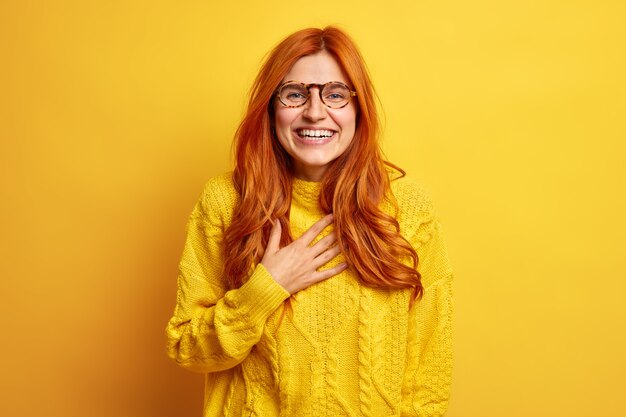 Portret uradowanej rudowłosej kobiety uśmiecha się szeroko, trzymając dłoń na piersi, uśmiecha się szeroko, wyraża dobre emocje, jest zadowolony, nosi przezroczyste okulary, swobodny sweter.