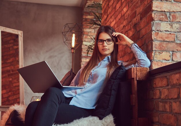 Portret uroczej brunetki bizneswoman w okularach i niebieskiej koszuli, pracującej online z laptopem i siedzącej na kanapie w pokoju o loftowym designie.