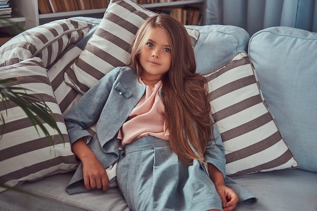 Bezpłatne zdjęcie portret uroczej dziewczynki z długimi brązowymi włosami i przeszywającym spojrzeniem, patrzącej w kamerę, leżącej samotnie na kanapie w domu.