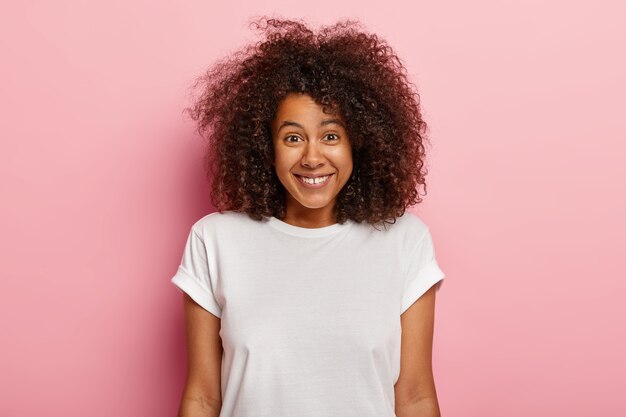 Portret uroczej młodej Afroameryki z bliska uśmiecha się pozytywnie, lubi zabawną scenę, jest rozbawiona, chichocze z zabawnego żartu, ma ciemne kręcone, obszerne włosy, nosi swobodny strój