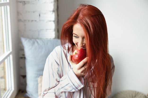 Portret uroczej, radosnej rudowłosej młodej Europejki w modnej piżamie w paski o szczęśliwym wyglądzie, gryzącej świeże dojrzałe czerwone jabłko, jedzącej owoce na śniadanie, siedzącej przy oknie w swoim pokoju