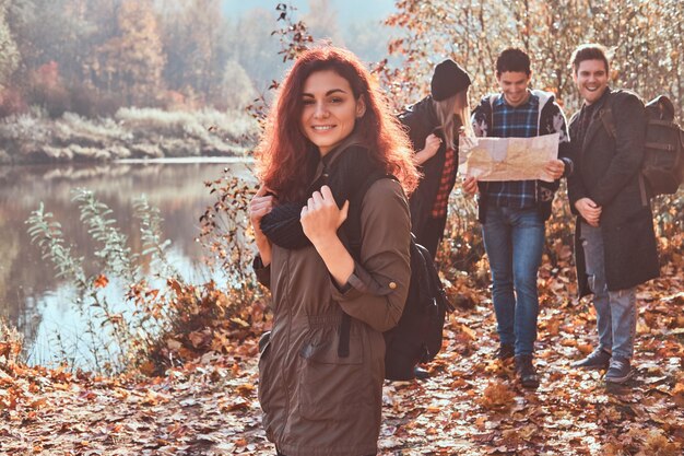 Portret uroczej rudej dziewczyny z plecakiem na pierwszym planie i grupą przyjaciół patrzących na mapę i planujących wędrówkę w tle w jesiennym lesie.