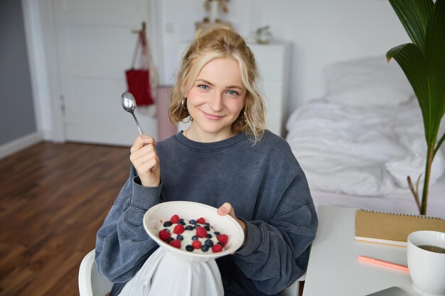 Bezpłatne zdjęcie portret uroczej uśmiechniętej blondynki jedzącej śniadanie w sypialni patrząc na miskę z kamerą