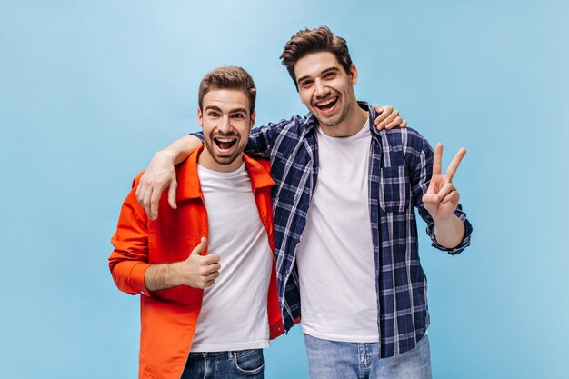 Portret uroczy brunet mężczyzn w białych koszulkach i dżinsach na na białym tle mężczyzna w pomarańczowej kurtce pokazuje uderzenie w górę i facet w kraciastej koszuli pokazuje znak pokoju