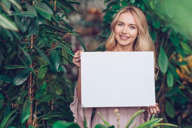 Portret uśmiechnięta blondynki młodej kobiety pozycja w rośliny pepinierze pokazuje białego pustego plakat
