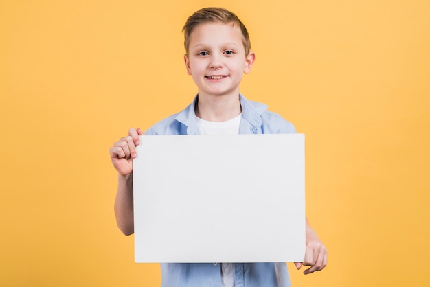 Bezpłatne zdjęcie portret uśmiechnięta chłopiec patrzeje kamera pokazuje białego pustego plakat przeciw żółtemu tłu