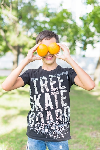 Portret uśmiechnięta chłopiec trzyma świeże całe pomarańcze nad jej oczami w parku
