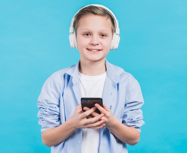 Portret uśmiechnięta chłopiec z białym hełmofonem na kierowniczym mienia smartphone w ręce przeciw błękitnemu tłu