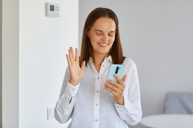 Portret uśmiechnięta ciemnowłosa młoda dorosła kobieta nosi białą koszulę, stojąc w domu z telefonem w rękach, po rozmowie wideo, machając ręką do aparatu urządzenia.