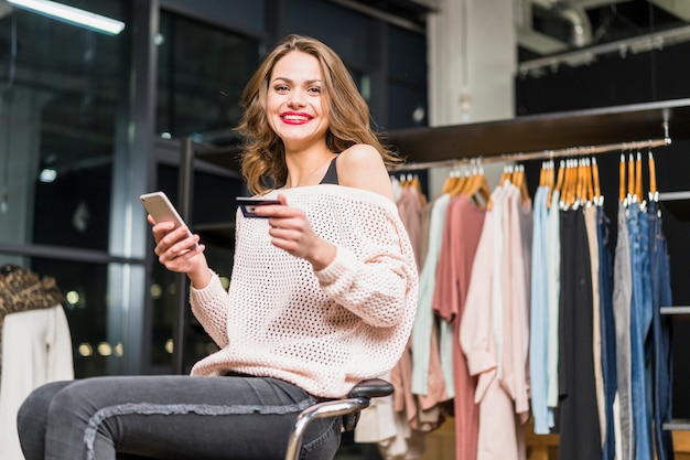 Portret uśmiechnięta kobieta siedzi w sklepie trzyma kartę kredytową i telefon komórkowy w ręku