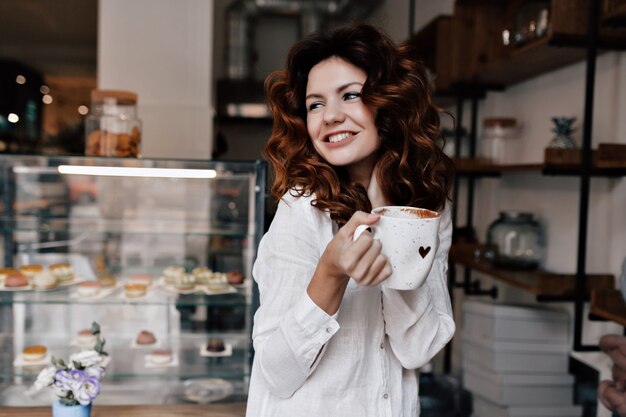 Portret uśmiechnięta młoda dama z filiżanką kawy stojąc przy kasie i czekając na klientów