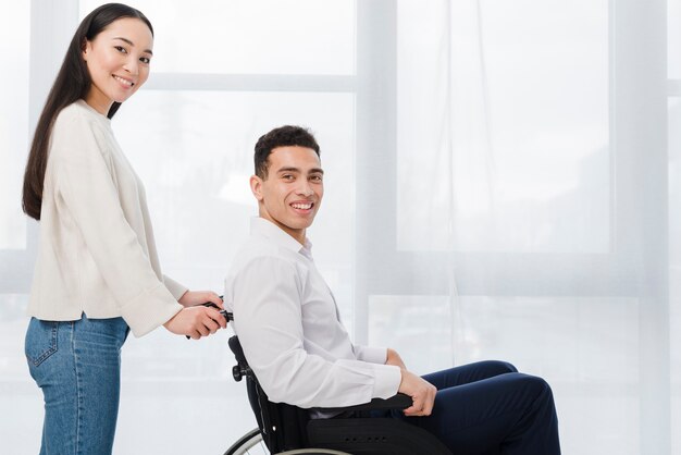 Portret uśmiechnięta młoda kobieta stoi za mężczyzna siedzi na wózku inwalidzkim, patrząc na kamery