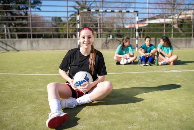 Portret uśmiechnięta nastoletnia dziewczyna piłki nożnej na polu. Szczęśliwa dziewczyna w sportowej odpoczynku na ziemi, patrząc na kamery, podczas gdy inne dziewczyny siedzą za nią, ciesząc się rozmową. Koncepcja aktywnego wypoczynku i sportów zespołowych