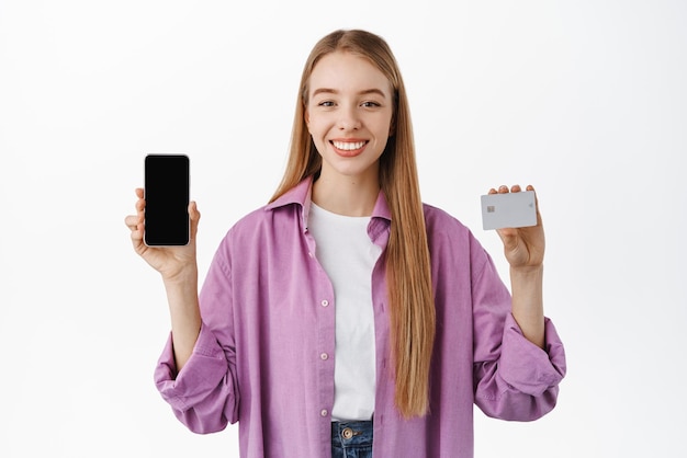 Portret uśmiechnięta nowoczesna dziewczyna pokazując pusty ekran smartfona i kartę kredytową wprowadzają zniżki na zakupy aplikacji w sklepie internetowym stojąc na białym tle Koncepcja reklamy
