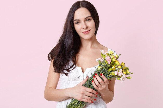 Portret uśmiechnięta szczera młoda modelka stojący na białym tle nad jasnoróżowym, trzymając w rękach kolorowe wiosenne kwiaty