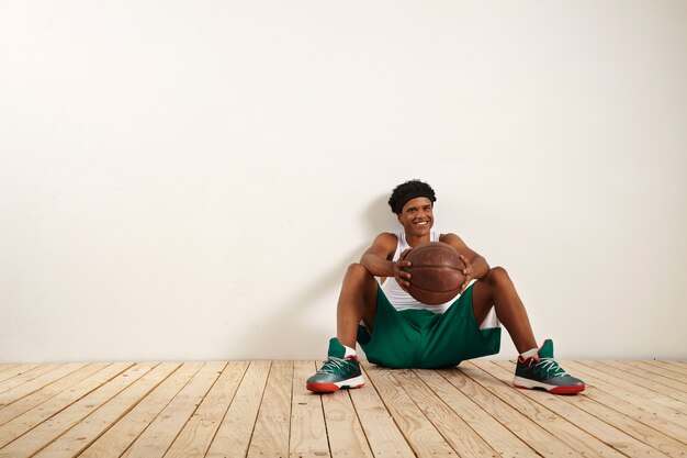 Portret uśmiechniętego młodego gracza siedzącego na drewnianej podłodze przy białej ścianie, trzymającego starą brązową piłkę do koszykówki