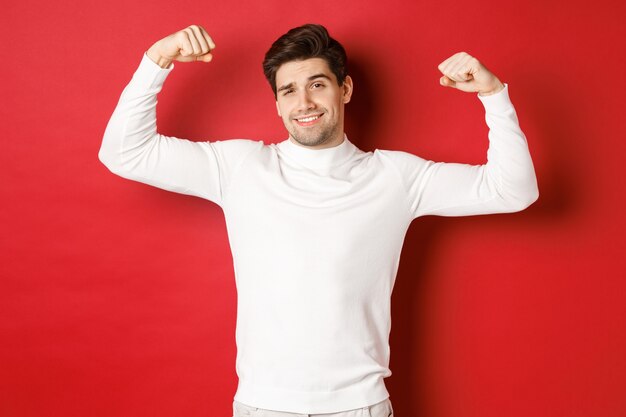 Portret uśmiechniętego przystojnego mężczyzny w białym swetrze napinającym bicepsy i chwalącym się pokazem siły ...