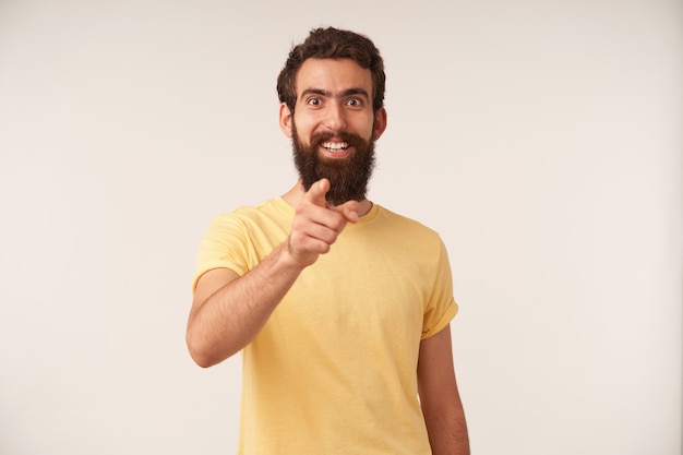 Bezpłatne zdjęcie portret uśmiechniętego przystojnego młodego faceta z brodą wskazuje palcem z przodu, patrząc na ciebie, ciesząc się, że zwraca uwagę na białej ścianie