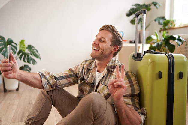 Bezpłatne zdjęcie portret uśmiechniętego przystojniaka pokazującego znak pokoju robi selfie na smartfonie siedzi z walizką
