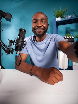 Portret uśmiechniętego słynnego influencera biorącego szerokokątny selfie w studiu nagrań podcastów, patrząc na kamerę przed profesjonalnym mikrofonem. pov ujęcie vlogera z mediów społecznościowych przy biurku.