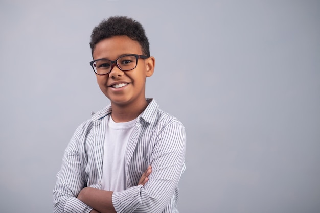 Portret uśmiechniętego, zadowolonego dziecka w okularach, stojącego z założonymi rękoma na białym tle