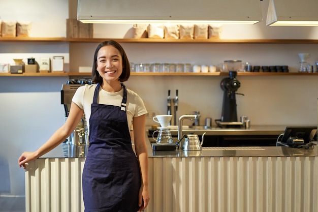 Portret uśmiechniętej azjatyckiej baristki w fartuchu stojącej przy ladzie z kawą pracującą w