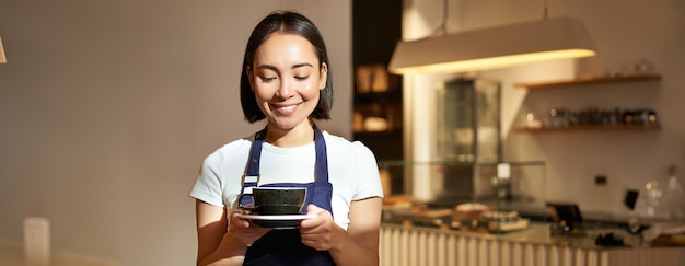 Portret uśmiechniętej azjatyckiej dziewczyny baristy w mundurze serwującej kawę kelnerka trzymająca filiżankę napoju