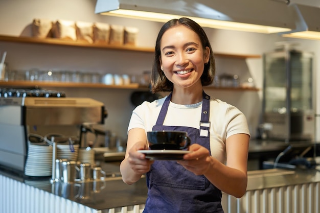 Portret uśmiechniętej azjatyckiej kobiety trzyma filiżankę kawy, przygotowuje napoje dla klientów w pracy w kawiarni i s