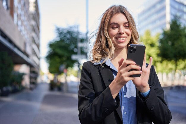 Portret uśmiechniętej bizneswoman korzystającej z telefonu komórkowego, stojąc na zewnątrz w pobliżu budynków biurowych